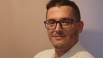 Ein Mann mit Brille trägt einen weißen Blazer und blickt freundlich in die Kamera.