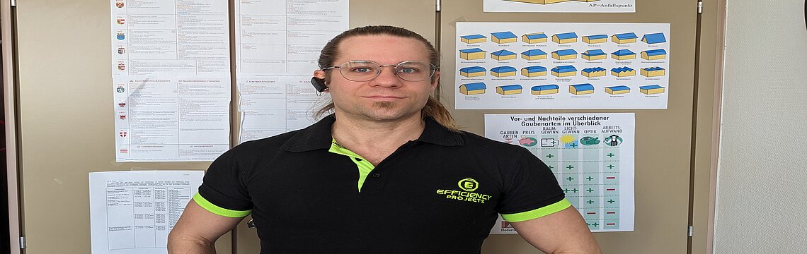 Ein Mann trägt ein schwarzes T-Shirt und steht vor einem Informationsbrett.