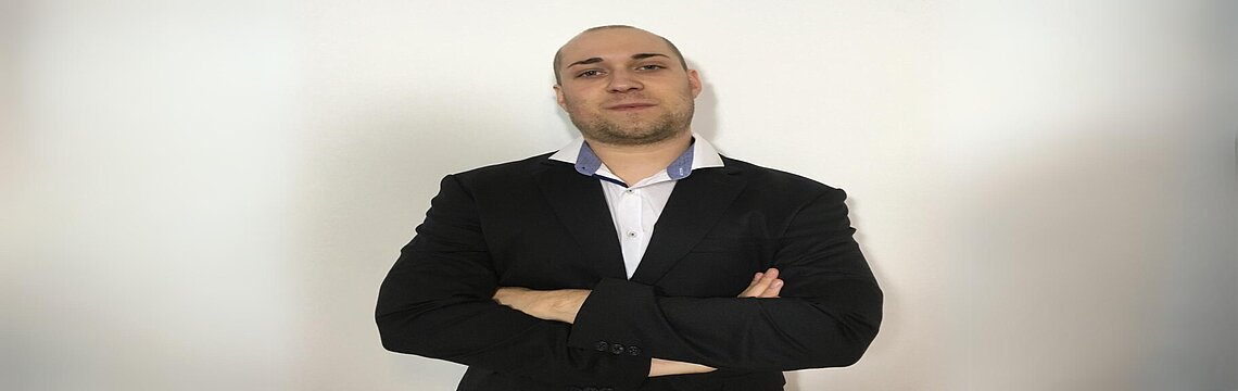 Ein Mann in Anzug steht mit verschränkten Armen und blickt selbstbewusst in die Kamera.