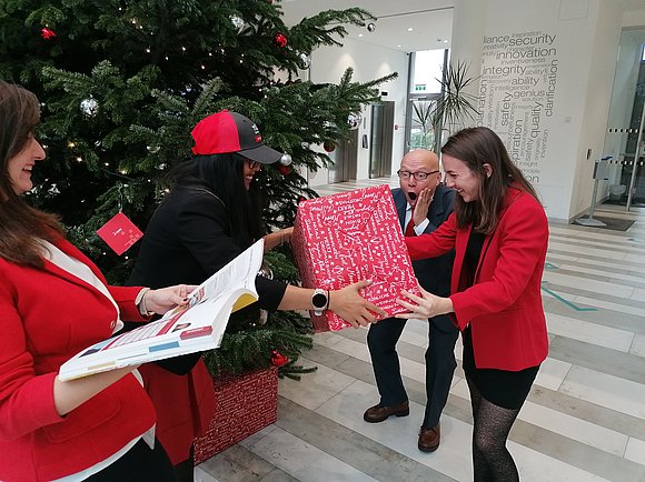 Zwei Damen überreichen einem Herren ein Weihnachtsgeschenk. Im Hintergrund ein Christbaum.