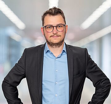 Ein Brillenträger in einem Anzug steht in einem Büro und strahlt Selbstbewusstsein und Professionalität aus.