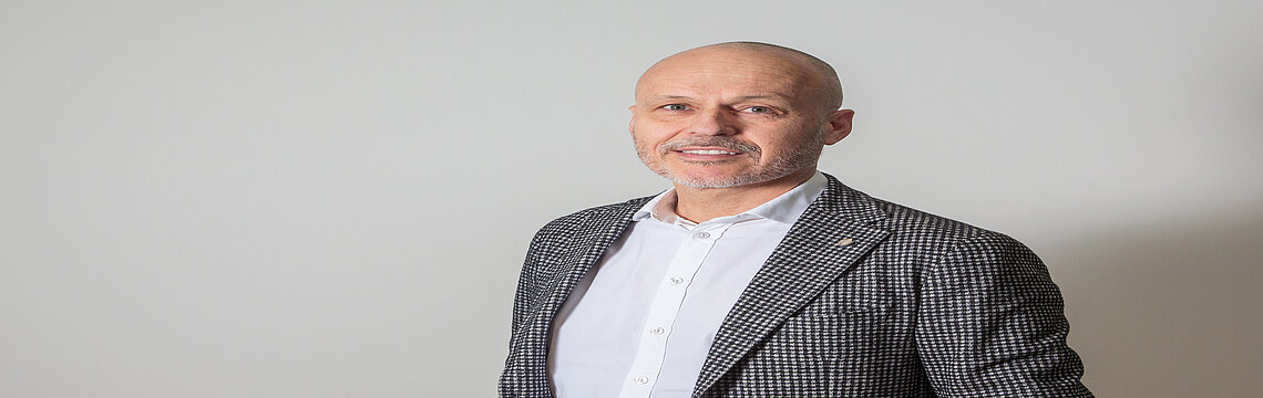 Ein glatzköpfiger Mann in formeller Kleidung vor einer weißen Wand.