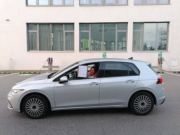 Dame mit FFP2 Maske sitzt in einem Auto und hält eingerahmte Zertifizierung in die Höhe.
