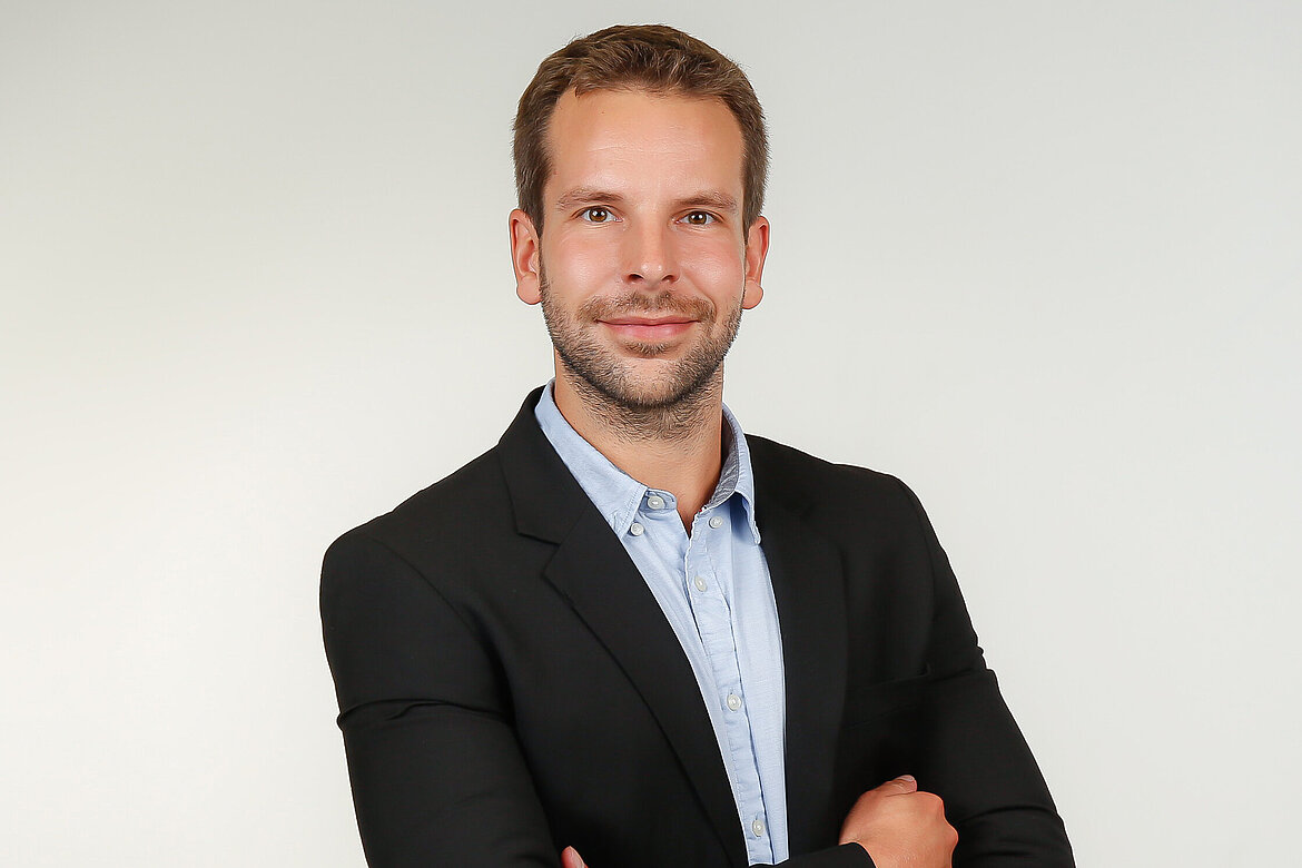 Ein Mann in Anzug und Krawatte steht mit verschränkten Armen da und lächelt in die Kamera.